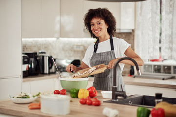 Attractive mixed race woman in apron standing in kitchen and putting vegetables in bowl. Dinner preparation.