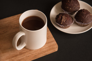 A white Cup of coffee stands on a wooden Board. Chocolate cupcakes are on the plate. Black background.