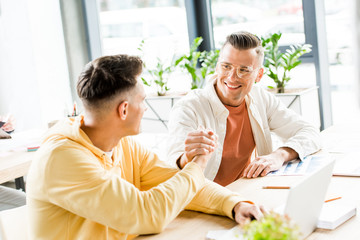 Fototapeta premium two young businessmen shaking hands while sitting together at workplace