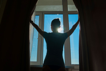 Fototapeta na wymiar Woman silhouette opening curtains and looking out of window in hotel room at morning