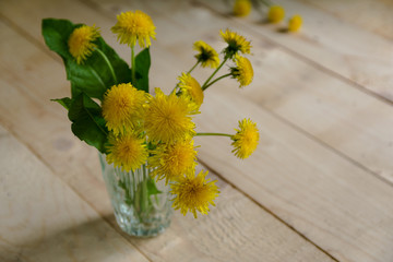 a bouquet of yellow dandelions in a transparent glass cup stands on a table of light wood