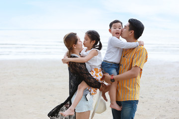 happy asian family dad and mom with their children boy and girl, parents carrying their children and kissing with love on sandy beach during sunny day..