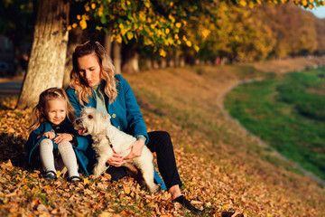 Naklejka na ściany i meble Happy mom with her child relaxing with white terrier dog on autumn leaves. Autumn season time. Fashion, family, autumn, lifestyle