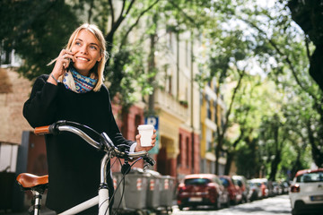 Fototapeta na wymiar Beautiful young woman posing with her bicycle and coffee while using phone