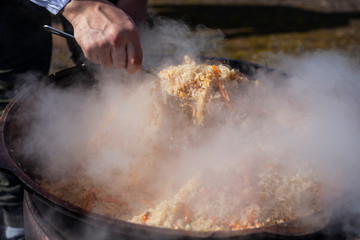 Cook cooks pilaf in a cauldron. Pilaf with vegetables and meat cooked over an open fire. Steam from hot food. A man interferes with rice.