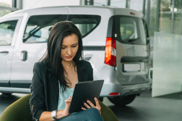 A businesswoman in a smart casual clothing sits in a car show on a background of a car and works with a tablet