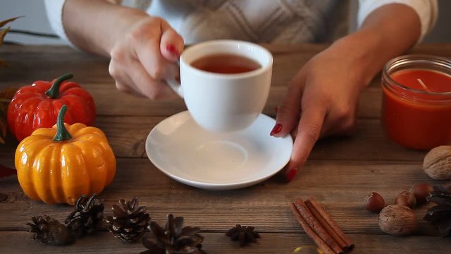 Woman's hand take and put white cup of tea on the old wooden table with pumpkin, leaves, spices on the background. Autumn concept.