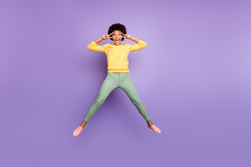 Fototapeta na wymiar Full body photo of amazed carefree afro american girl jump have fun on autumn spring holidays make v-signs wear yellow pullover green pants trousers isolated over violet purple color background