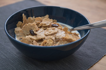 A dark blue colored bowl of delicious corn flake cereals with raisins, milk and a spoon beside a wooden surface.
