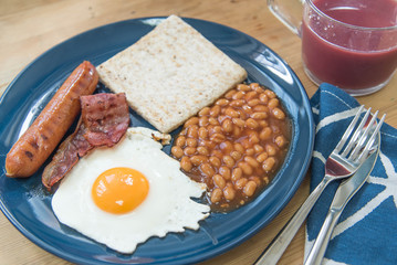 Full English breakfast with fried eggs, sausages, bacon, baked beans, toasts and mixed berry juice on a wooden surface
