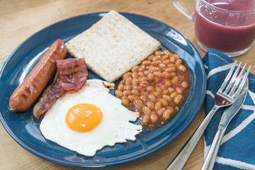Full English breakfast with fried eggs, sausages, bacon, baked beans, toasts and mixed berry juice on a wooden surface