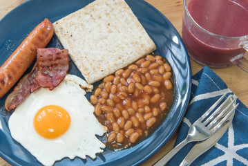 Full English breakfast with fried eggs, sausages, bacon, baked beans, toasts and mixed berry juice on a wooden surface