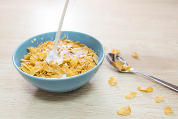 Milk pouring into a sky blue colored bowl of delicious corn flake cereals on a wooden surface