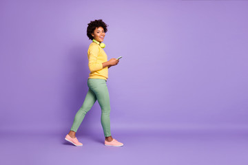 Full length body size photo of side profile walking smiling toothy girl going side profile holding telephone wearing yellow sweater green pants trousers isolated over violet pastel color background