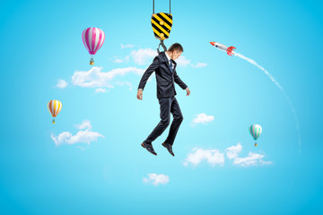 Businessman hanging on construction crane hook with hot air balloons and silver red space rocket in...