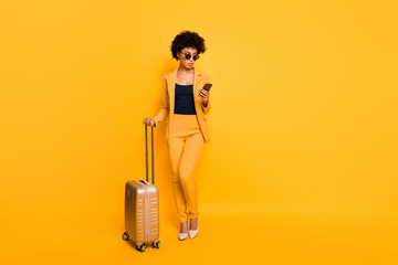 Full length photo of focused brown curly hair girl use cellphone search taxi hold bags tourism concept have abroad vacation wear style jacket pants high-heels isolated over yellow color background