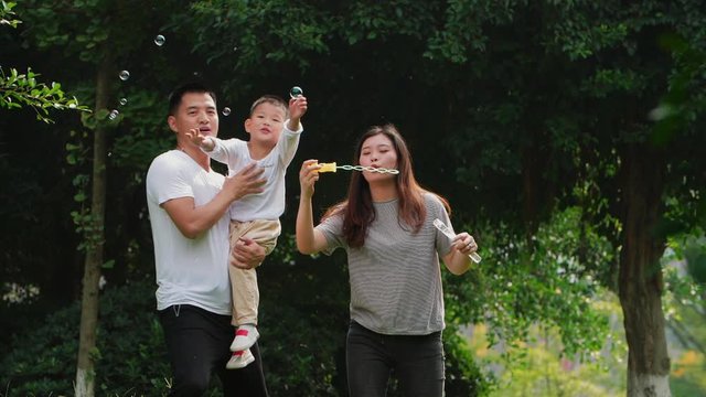 Happy asian family having good time outdoor in the sunny park father holding his son catching soap bubble in air parents playing with little boy joyful childhood