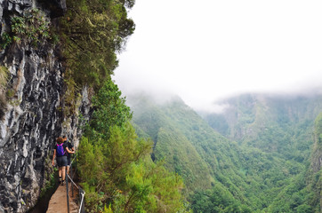 Hikers walking on a narrow path on the edge of the rock during Levada do Caldeirao Verde Trail. Misty green mountains in background. Dangerous hiking. Portuguese tourist attraction. Fog, foggy