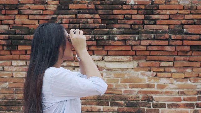 Side view of pretty Asian girl taking photos of tourist destinations standing next to an old brick wall - Sightseeing woman exploring historic city while travelling solo - social influencer concept