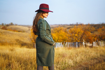 Beautiful young stylish girl in a coat walks in the autumn in the park. The girl is dressed in a green coat and a red hat. Beautiful evening. Autumn fashion. Lifestyle. High fashion portrait. 