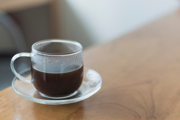 Hot black coffee in a transparent glass cup on a wooden table and blurred home or office background, shallow depth of field.