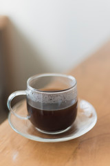 Hot black coffee in a transparent glass cup on a wooden table and blurred home or office background, shallow depth of field.