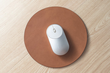 White and silver wireless mouse on a light brown round leather mouse pad on a wooden surface.