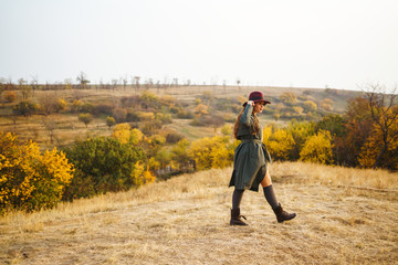 Beautiful young stylish girl in a coat walks in the autumn in the park. The girl is dressed in a green coat and a red hat. Beautiful evening. Autumn fashion. Lifestyle. High fashion portrait. 