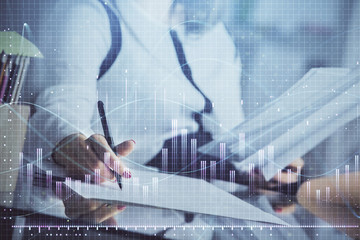 A woman hands writing information about stock market in notepad. Forex chart holograms in front. Concept of research.