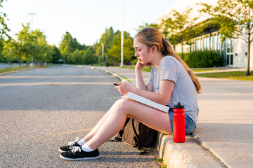 Depressed/Sad teen girl sitting on a curb in front of a high school during sunset while sitting...