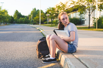 Depressed/Sad teen girl sitting on a curb in front of a high school during sunset while sitting next to a backpack and holding binders.