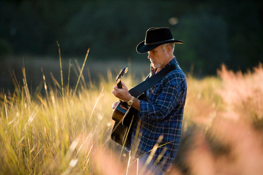 Mature man wearing a hat and strumming an acoustic guitar outdoors.