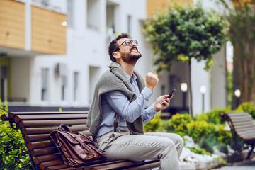 Fototapeta premium Successful handsome fashionable caucasian businessman sitting on bench in park, holding smart phone and rejoicing for achievement. Next to him is leather handbag.
