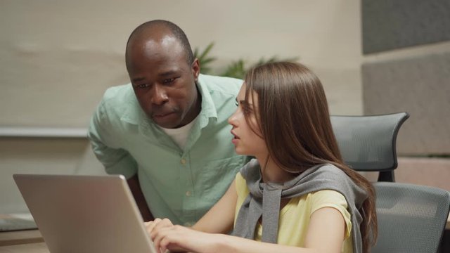 Camera panning right to concentrated African American man and young woman checking notes in notepad and using laptop while working on project in cubicle together