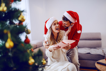 Amazed cute Caucasian blonde woman sitting on sofa in living room, hugging her boyfriend and...
