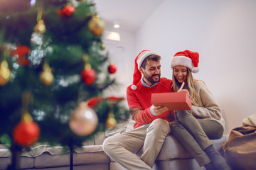 Handsome smiling bearded caucasian man giving christmas present to his adorable girlfriend. Both...