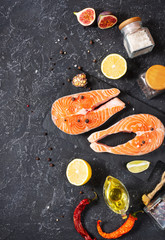 Two fresh raw salmon steaks with salt, peppers, lemon on black stone background.