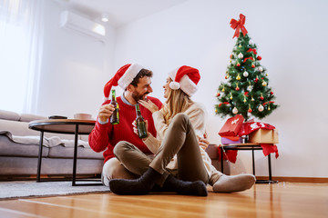 Adorable good-looking Caucasian couple with santa hats on heads sitting on floor in living room,cuddling and holding beer. In background is christmas tree with presents.
