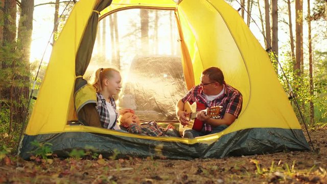 Happy father playing guitar to family in tent. Cheerful middle aged man playing acoustic guitar to smiling family sitting in yellow tent at camping