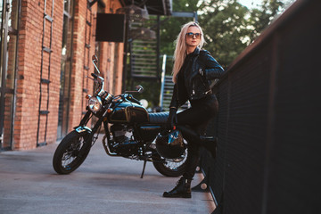 Plakat Blond female biker is posing for photographer next to black, shiny chopper.
