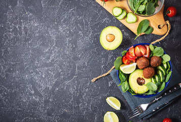 Salad with avocado, falafel, cucumber, cherry tomato and spinach on black stone background. Healthy vegan lunch bowl.