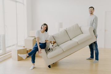 Happy couple move furniture in their new modern home, carry sofa with pet, pose in spacious room,...
