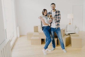 Fototapeta na wymiar Young couple in love stand closely to each other, embrace and holds favourite dog, pose in empty room, surrounded with cardboard boxes, make family portrait in new apartment. Moving Day concept