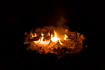 A campfire at night showing logs, flames and burning embers with dark background.