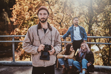 Attractive hipster in glasses is posing for photographer with photo camera while his friends are resting at background.