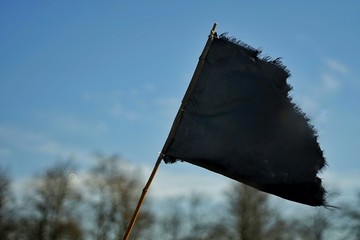 tilted black flag on a pole, against a blue sky and trees