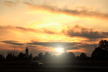 Pomarańczowo złoty zachód słońca nad wioską, wierza kościoła, samochód.