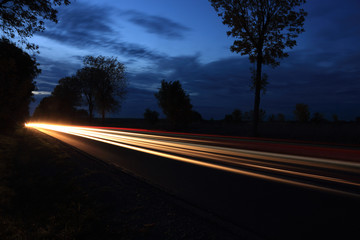 Światła samochodów w nocy na drodze między polami o zmierzchu.