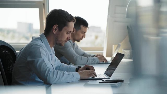 Two young intelligent men in white shirts are sitting in front of laptops in a bright office in front of a large window. One of the men uses a mobile phone, responds to the message.