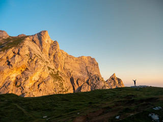Hiker standing on the edge of grassy hill in the Alps raises her arms in victory